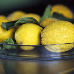 Natural, mediterranean lemons with leaves in glass dish on stainless steel counter